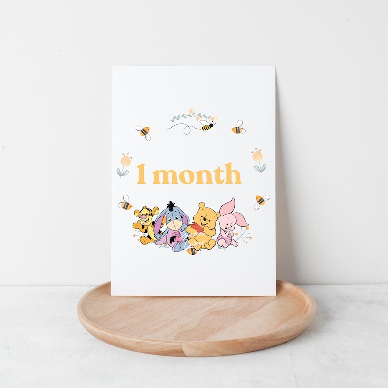 May include: A white card with a yellow background featuring Winnie the Pooh, Piglet, Tigger, and Eeyore. The card says "1 month" in yellow text. The card is surrounded by bees and flowers.