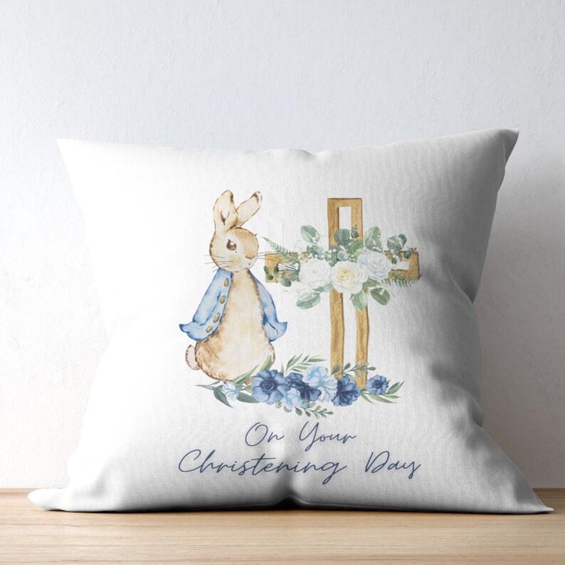 Peut inclure: Un oreiller blanc avec une illustration aquarelle de Lapin de P&acirc;ques debout &agrave; c&ocirc;t&eacute; d'une croix en bois d&eacute;cor&eacute;e de roses blanches et de fleurs bleues. Le texte "On Your Christening Day" est &eacute;crit sous l'illustration.