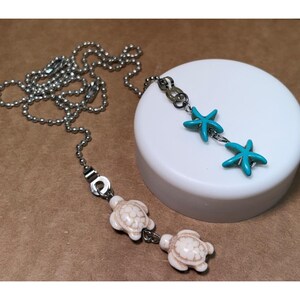 May include: A silver-toned ball chain necklace featuring two distinct charms. One charm consists of two turquoise-colored starfish, while the other charm features two off-white sea turtle figures with detailed shell patterns connected by small metal rings.