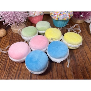 May include: A collection of colorful, plush macaron ornaments in shades of pink, blue, yellow, and green. Each ornament has a white, fluffy filling and a clear ribbon for hanging. The macarons are arranged on a wooden surface.
