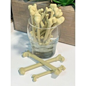 May include: A glass filled with multiple beige plastic bone-shaped stir sticks. Three additional bone-shaped stir sticks are lying on a white surface.