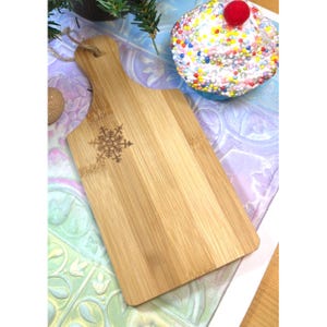 May include: A rectangular bamboo cutting board with a handle and a snowflake design. The board is light brown with a natural wood grain. A decorative cupcake with colorful sprinkles and a red cherry is in the background.