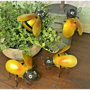 May include: Three metal bee figurines featuring black heads, yellow and orange wings, and thin wire legs. Each bee has a rounded body with black stripes. The bees are positioned around a green plant in a wooden bowl and a gray metal planter.