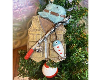 Puede incluir: Un adorno navideño con temática de pesca. El adorno presenta un sombrero de pescador azul claro, un chaleco de pesca color canela, una caña de pescar y un señuelo. Una boya roja y blanca cuelga debajo. El adorno está decorado con agujas de pino verdes falsas.