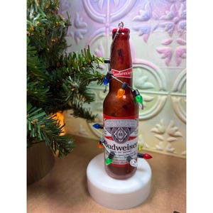 May include: A brown glass Budweiser beer bottle ornament wrapped in a string of small, multi-colored LED Christmas lights. The bottle features the classic red, white, and blue Budweiser label with the brand name and logo clearly visible on the front.