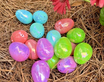 May include: A collection of colorful, iridescent plastic Easter eggs in shades of blue, pink, green, and purple. The eggs are scattered on a bed of brown pine needles, with a red flower in the background. A festive and decorative item.