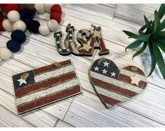 May include: Three wooden ornaments with a patriotic theme. The ornaments are shaped like a heart, a rectangle, and the letters USA. Each ornament features a red, white, and blue American flag design with white stars.