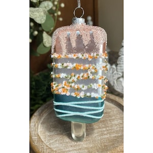 May include: A glass ornament shaped like an ice cream popsicle with a pink and white frosting design and blue and white stripes. The ornament has a silver hanger.