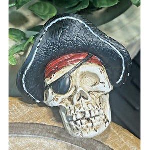 May include: A white skull wearing a black pirate hat with a red and white bandana and an eye patch. The skull has a detailed face with teeth and a prominent nose.