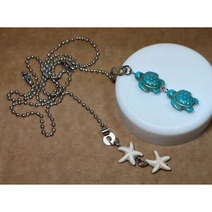 May include: A silver-toned ball chain necklace featuring two turquoise-colored sea turtle charms and two white starfish charms. The charms are attached to the metal chain, which has a lobster claw clasp, creating a nautical-themed accessory.