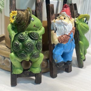 May include: Three decorative garden ornaments featuring a green turtle with a sunflower, a gnome with a white beard in blue overalls, and a green frog, each climbing a separate brown wooden ladder with a hook at the top for hanging display.