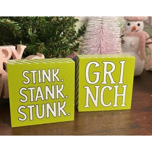 May include: Two square lime green wooden blocks with white diagonal stripes on the sides. The left block features the white text STINK. STANK. STUNK. and the right block features the white text GRINCH in a bold, sans-serif font style.