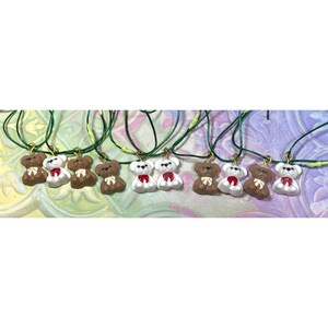 May include: A collection of small bear-shaped pendants hanging from thin green cords. The bears are rendered in either solid white or brown, each featuring a small painted bow in either red or white around the neck and a gold-toned metal jump ring.