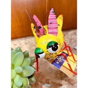 May include: A metal insect figurine with a bright yellow body, pink and yellow wings, and large green and white eyes. The insect has thin red legs and a single antenna. A small rectangular tag with the text "the spring" is attached to the base.