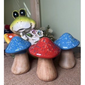 May include: Three ceramic mushroom figurines with speckled blue, red, and brown paint. The mushrooms have brown stems and are sitting on a brown surface.