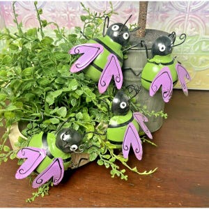 May include: Four metal bee ornaments featuring lime green bodies with black stripes, black heads with white eyes, and lavender wings with black swirl details. These decorative insects are positioned among green foliage.