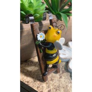 May include: A whimsical figurine of a bee climbing a miniature wooden ladder. The bee is yellow and black, holding a small bouquet of blue and white flowers. The ladder has a brown finish with black metal hooks.