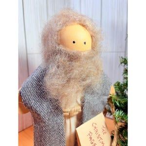 May include: A handmade Christmas figure with a wooden head, a long, light brown beard, and a gray herringbone patterned coat. The figure has a beige robe and holds a tag that reads "The Christmas Page".