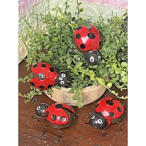 May include: Four metal ladybug figurines featuring bright red wings with black spots and black heads with white eyes. Three of the ladybugs are positioned on a wooden surface, while one is nestled within a green plant in a round wooden bowl.