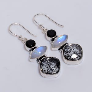 May include: Silver dangle earrings with a black onyx stone, a pearlescent moonstone, and a square-cut black tourmaline stone with inclusions. The earrings have a hook closure.