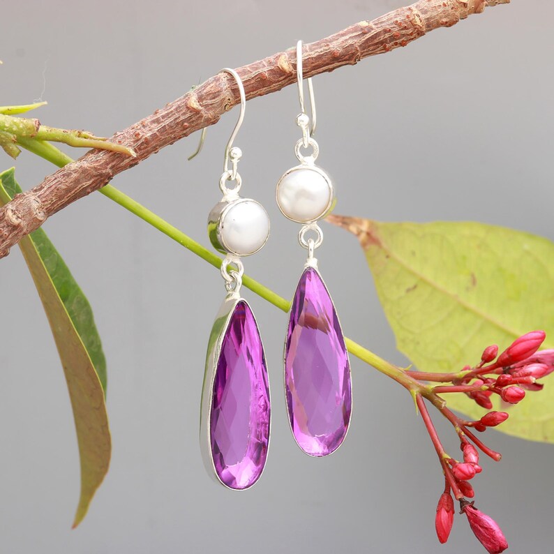 May include: A pair of silver dangle earrings featuring a teardrop-shaped purple gemstone and a small white pearl accent.