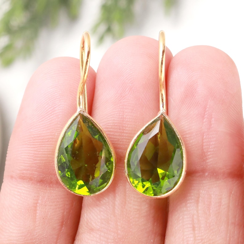 Puede incluir: Un par de pendientes colgantes dorados con piedras preciosas en forma de pera, facetadas, de color verde oliva. Los pendientes tienen un dise&ntilde;o sencillo y elegante, con cierre de gancho. Las piedras son de un verde transl&uacute;cido vibrante.