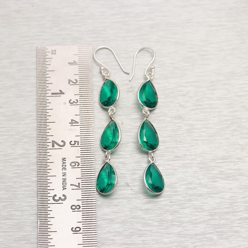 Op de afbeelding: Een paar lange oorbellen met elk drie smaragdgroene, druppelvormige stenen, gezet in zilver. De oorbellen worden getoond naast een liniaal.