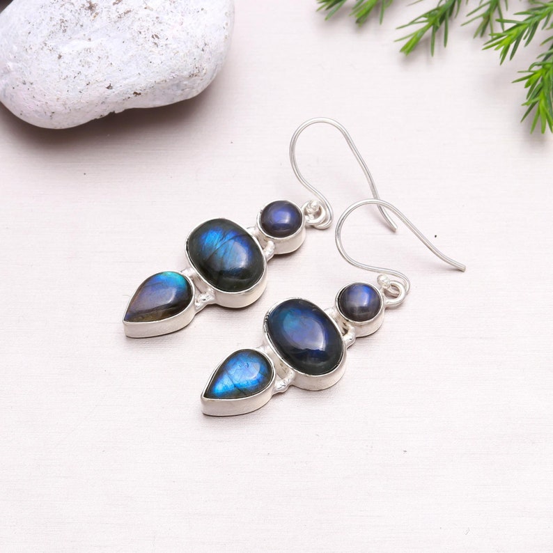May include: Silver dangle earrings with three iridescent blue labradorite gemstones. The earrings have a teardrop, oval, and round shape.