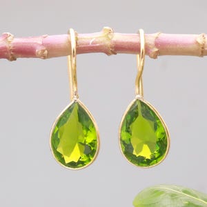 Puede incluir: Un par de pendientes dorados con gemas verdes claras facetadas en forma de pera. Los pendientes cuelgan de un fino gancho dorado curvado. Las gemas son de un color verde brillante y los pendientes se muestran sobre un fondo suave y neutro.
