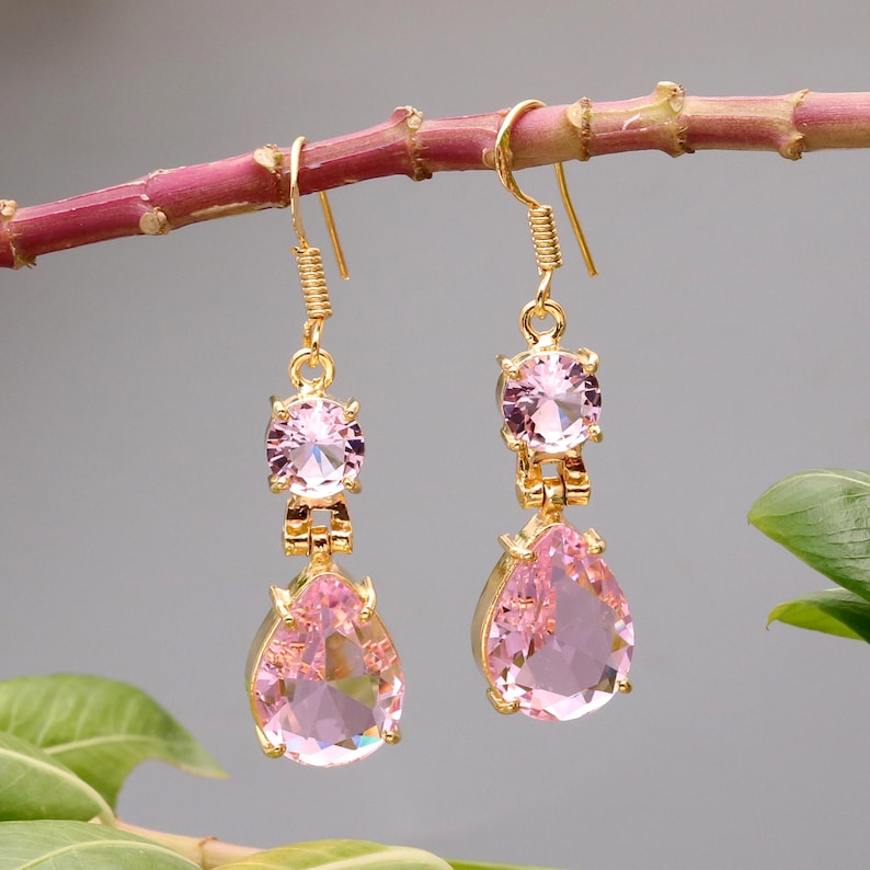 May include: A pair of gold-colored drop earrings with pink gemstones. Each earring features a small round gem at the top, a connecting piece, and a larger, pear-shaped pink gem at the bottom. The earrings are hanging from a pink branch.