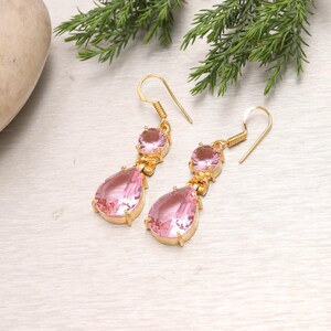 May include: A pair of gold-colored drop earrings featuring pink gemstones. Each earring has a round gemstone at the top and a teardrop-shaped gemstone below. The earrings are displayed on a light-colored surface with green foliage in the background.
