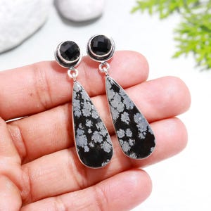 Könnte beinhalten: Ein Paar silberfarbene Hängeohrringe. Jeder Ohrring hat einen schwarzen, runden Stein oben und einen tropfenförmigen Stein mit schwarzem Grund und weißen schneeflockenartigen Mustern. Die Ohrringe werden in einer Hand gehalten.