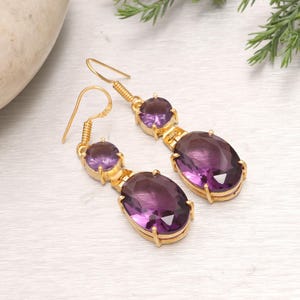 Peut inclure: Une paire de boucles d'oreilles pendantes dorées ornées de deux pierres ovales violettes à facettes. La pierre plus petite est placée au-dessus d'une pierre ovale plus grande, fixées dans des montures en métal doré avec un crochet.