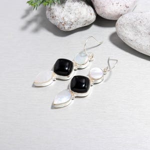 May include: A pair of silver earrings with a dangling design. Each earring features a black square stone, a white round stone, and a white teardrop-shaped stone. The earrings are displayed on a silver surface.
