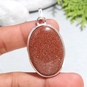 May include: A silver pendant with a large oval-shaped red sunstone gemstone. The stone has a sparkly, glittery texture.