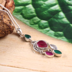 Può includere: Una collana con ciondolo in argento con un design di pietre preziose rosse, bianche e verdi. Il ciondolo presenta una grande pietra preziosa rossa al centro, circondata da pietre preziose bianche e verdi più piccole.
