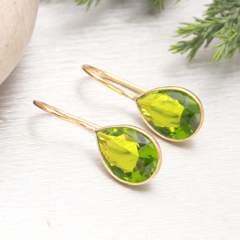 Puede incluir: Un par de pendientes colgantes dorados con piedras preciosas verde claro facetadas en forma de pera. Los pendientes tienen un dise&ntilde;o sencillo y elegante, con las piedras engastadas en un bisel dorado.