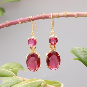 May include: Gold-toned dangle earrings with two ruby-red gemstones. The top stone is round, and the bottom stone is an oval shape. The earrings are hanging from a pink branch against a gray background.