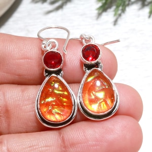 Op de afbeelding: Zilveren oorhangers met een kleine, ronde, rode edelsteen en een grotere, druppelvormige, oranje edelsteen. De oranje edelstenen hebben een iriserende glans. Een opvallend accessoire.