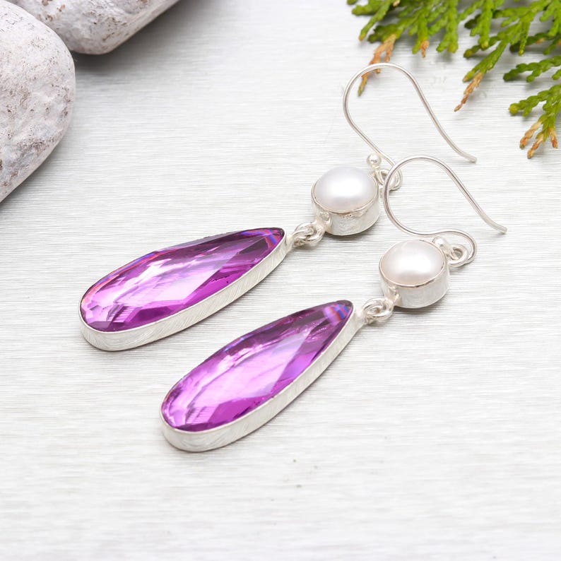 May include: A pair of dangle earrings featuring a teardrop-shaped purple gemstone and a white pearl accent. The earrings are made of sterling silver.