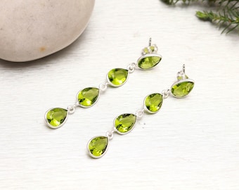 Puede incluir: Un par de pendientes colgantes de plata con tres piedras de peridoto verde claro en forma de pera. Los pendientes se muestran sobre una superficie blanca, con una piedra natural y vegetación en el fondo.