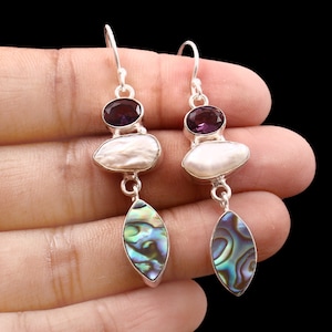 Op de afbeelding: Zilveren oorhangers met een paarse edelsteen, een witte parel en een blauwe abalone-schelp.