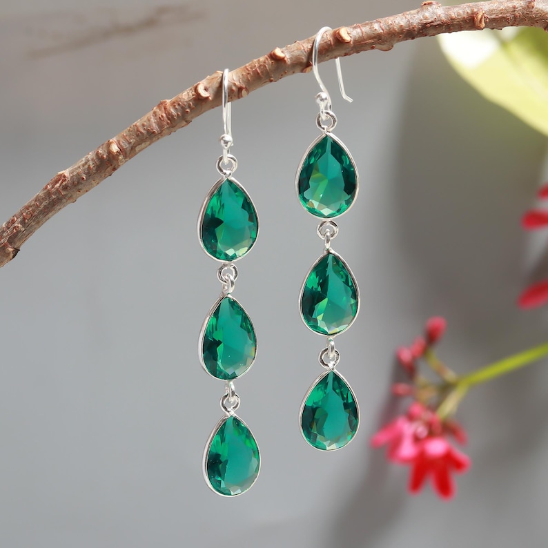 Op de afbeelding: Elegante zilveren oorbellen met drie smaragdgroene, druppelvormige stenen. De oorbellen hangen aan een zilveren haak en zijn verbonden door kleine zilveren schakels. De oorbellen worden gepresenteerd op een bruine tak tegen een zachte grijze achtergrond.