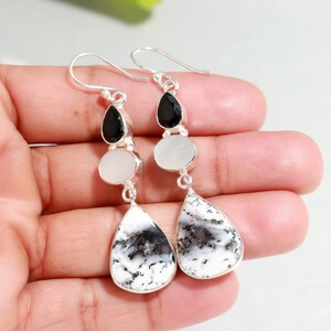 May include: A pair of dangle earrings with a silver hook. Each earring features a black gemstone, a white circular stone, and a teardrop-shaped white stone with black and gray patterns. The earrings are held in a hand.