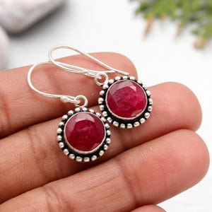 May include: A pair of silver dangle earrings with red ruby gemstones set in a silver bezel. The earrings have a simple hook closure.