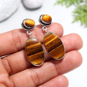 May include: A pair of silver earrings with oval-shaped tiger's eye gemstones. The earrings have a smaller round tiger's eye gemstone at the top of the oval.