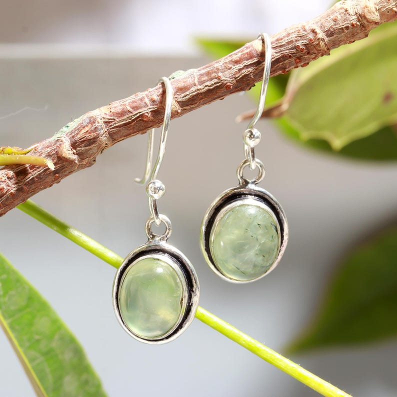 May include: A pair of silver dangle earrings with oval-shaped green gemstones. The earrings have a simple design and are perfect for everyday wear.