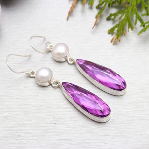 May include: A pair of dangle earrings featuring a pear-shaped purple gemstone and a white pearl accent. The earrings are made of silver.