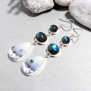 May include: A pair of silver earrings featuring a teardrop-shaped white stone with black inclusions, connected to two blue-green stones. The earrings have a hook closure and are set against a light gray background.