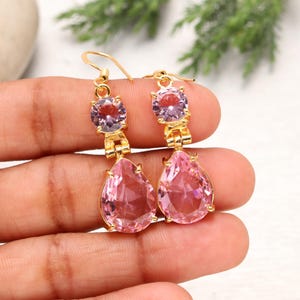 May include: A pair of gold-colored drop earrings. Each earring features a small, round, light purple gemstone at the top, connected to a larger, pear-shaped, pink gemstone. The earrings are held up by a gold-colored hook.